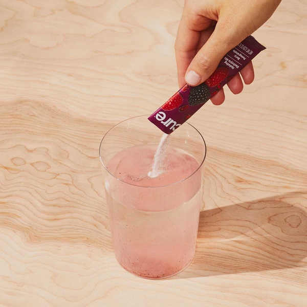 A close-up, high-angle shot of a hand pouring a berry-flavored electrolyte mix powder packet into a glass of water on a light wood surface.